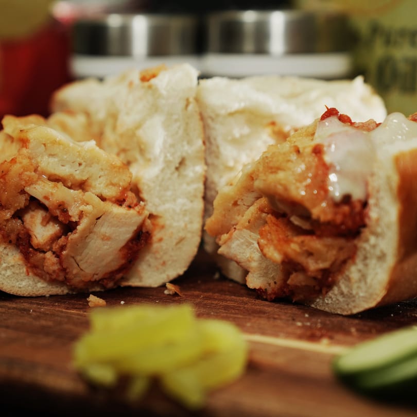 A chicken cheesesteak sandwich, cut in half, on a wooden board with pickled peppers.