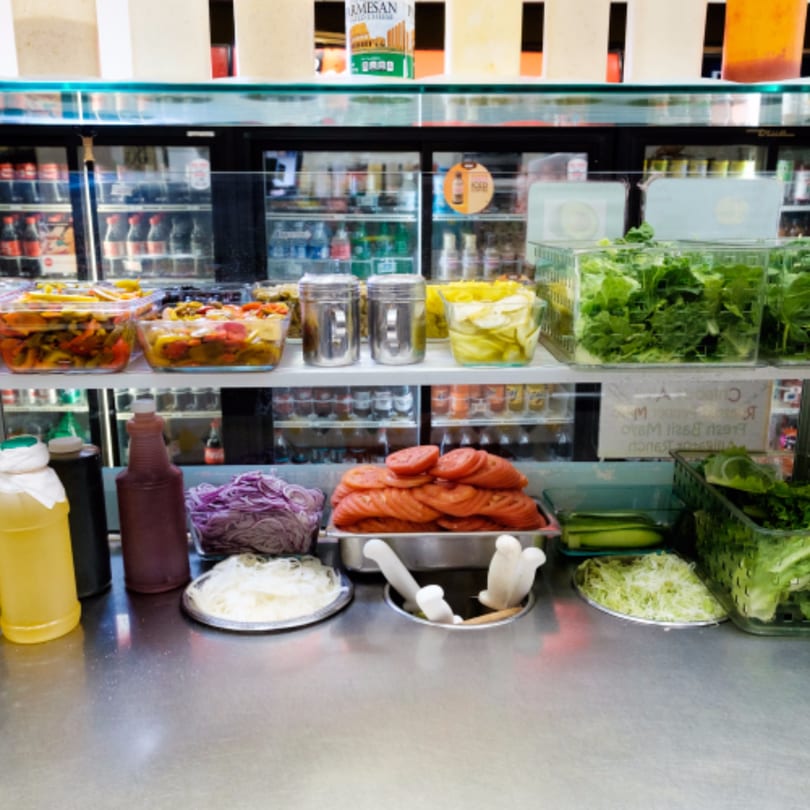 A sandwich bar with various fresh toppings like tomatoes, lettuce, onions, and pickled vegetables.