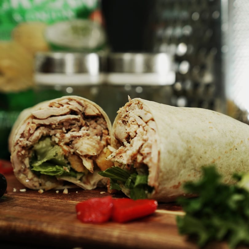 Two halves of a chicken and lettuce wrap on a wooden board with red peppers.
