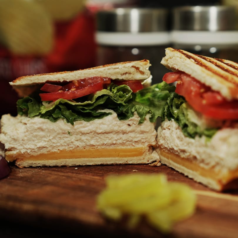 A grilled tuna melt sandwich with lettuce, tomato, and cheese, cut in half on a wooden board.
