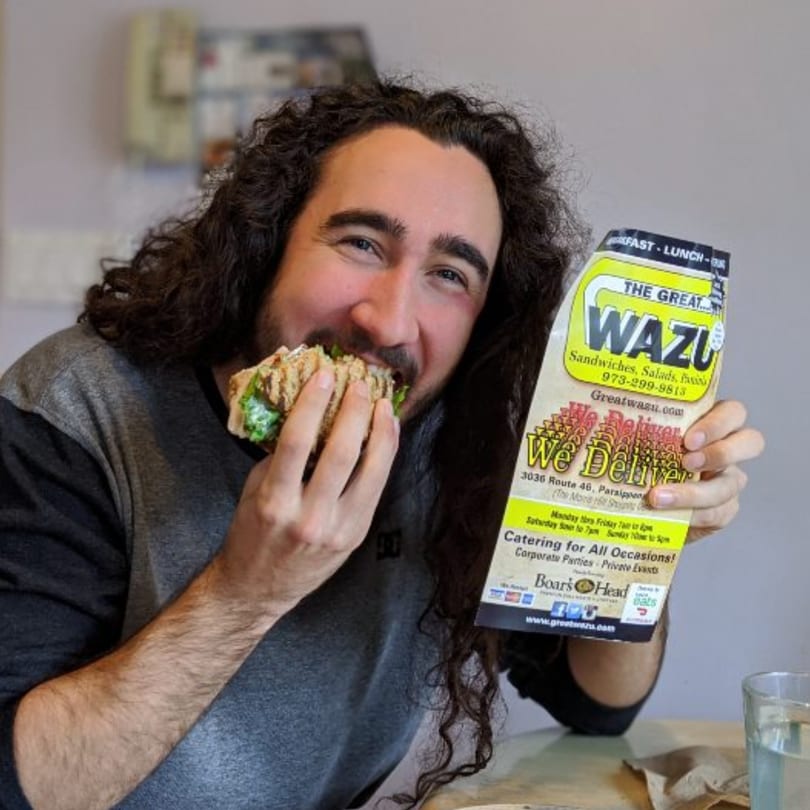 A man with long curly hair happily bites into a sandwich while holding a Great Wazu menu.