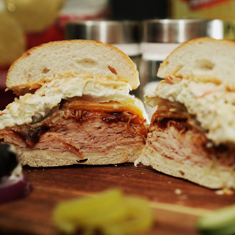 A Wazu sandwich, cut in half, revealing layers of meat, sauce, and coleslaw on a wooden board.