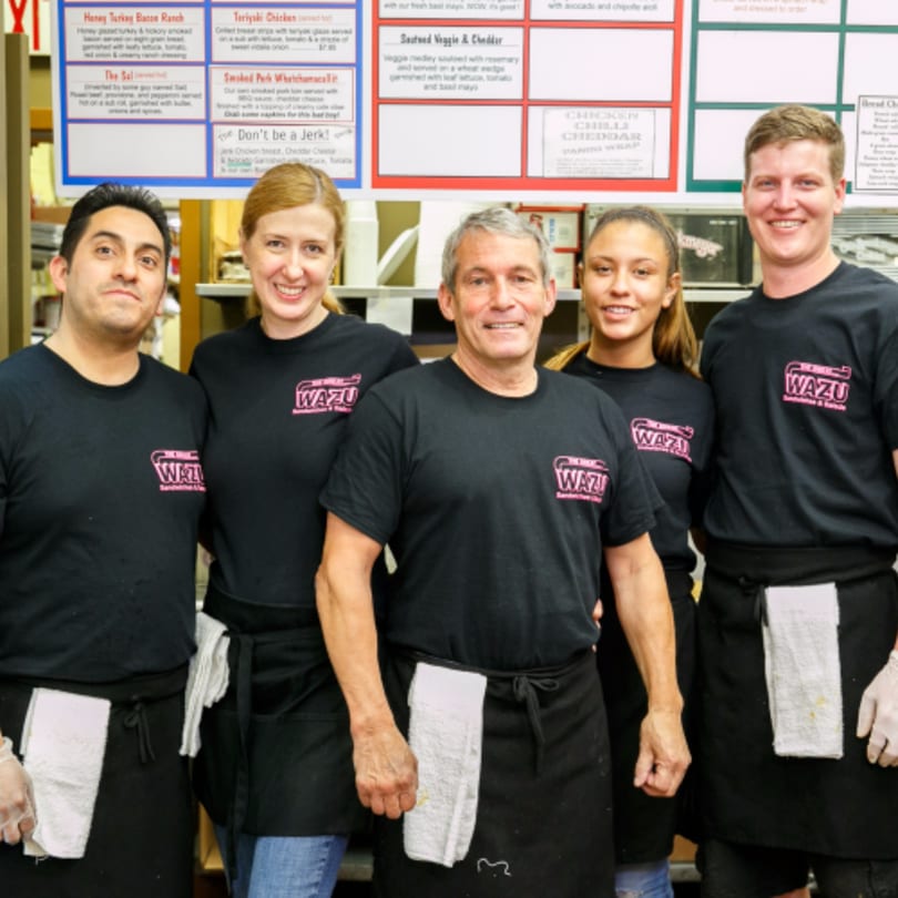 Five smiling Great Wazu sandwich shop employees in black shirts and aprons stand together.
