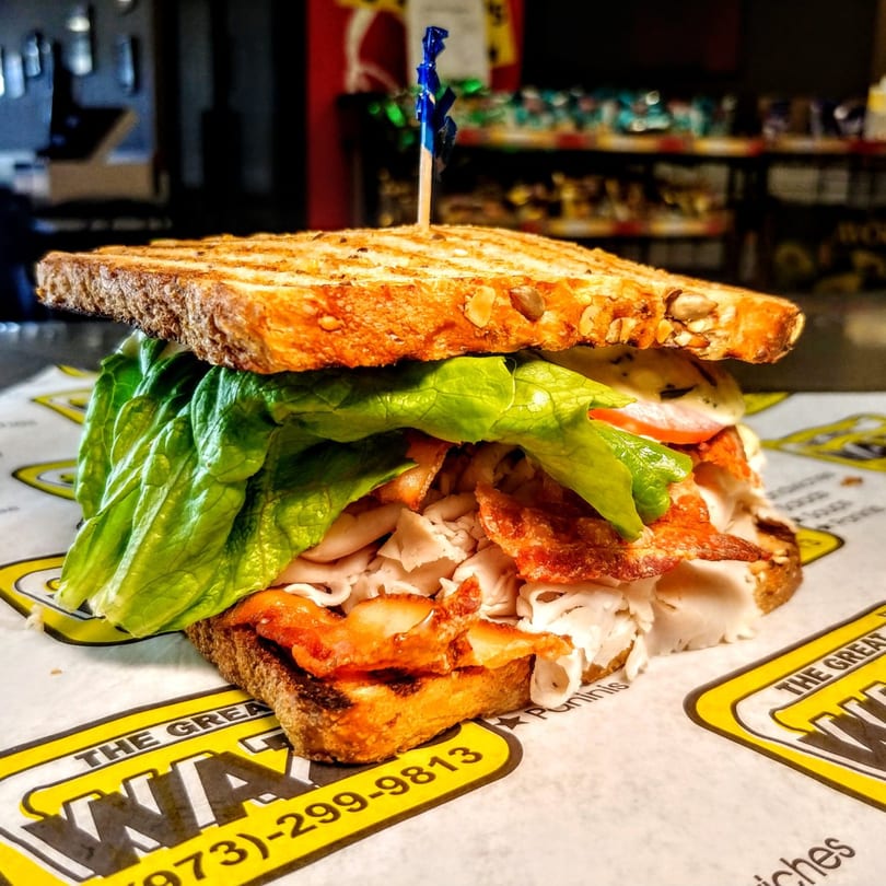 A grilled turkey and bacon sandwich with lettuce and tomato on branded deli paper.